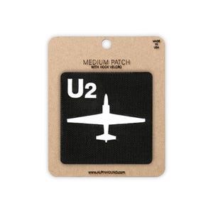 May include: A medium-sized black patch with a white U2 logo and aircraft design, attached to a cardboard backing. The text "MEDIUM PATCH WITH HOOK VELCRO" and "MADE IN USA" are visible. The website address www.alpinhound.com is also printed on the backing.