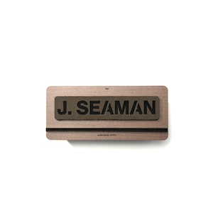 May include: A wooden name tag holder with a green fabric patch displaying "J. SEAMAN" in black capital letters. The holder features a black elastic band and the text "ALPHAHOUND SUPPLY" is printed below the band.
