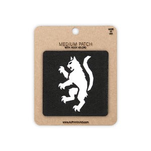 May include: A square black patch featuring a white graphic of a stylized cat in a rampant pose. The patch is mounted on a rectangular tan cardboard backing labeled MEDIUM PATCH WITH HOOK VELCRO and WWW.ALPINHOUND.COM with MADE IN USA text.