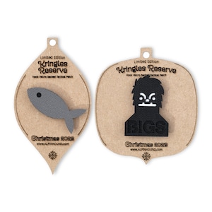 May include: Two Christmas ornaments shaped like teardrops with a brown background. The ornament on the left features a gray fish with a black eye. The ornament on the right features a black yeti with a white face and the word "BIGS" in white letters. Both ornaments have the text "Limited Edition Kringles Reserve Hook Velcro backed Tactical Patch Christmas 2023 www.ALPINHOUND.com".