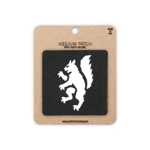 May include: A black square patch featuring a white silhouette of a squirrel in a rampant pose. The patch is mounted on a tan cardboard backing labeled MEDIUM PATCH WITH HOOK VELCRO, MADE IN USA, and the website address WWW.ALPINHOUND.COM.