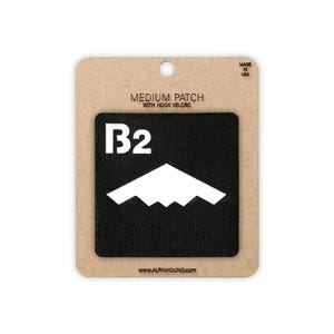 May include: A medium-sized black patch with white text and a mountain-like graphic. The patch reads "B2" and "www.alpinhound.com" on a cardboard backing. The patch is labeled "Medium Patch with Hook Velcro" and "Made in USA".
