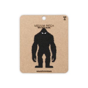 May include: A black fabric patch shaped like a silhouette of a Bigfoot creature with white eyes, mounted on a rectangular tan cardboard backing. The text reads MEDIUM PATCH WITH HOOK VELCRO, MADE IN USA, and www.ALPINHOUND.COM.