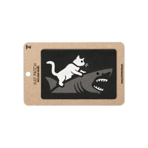May include: A rectangular black patch featuring a white embroidered graphic of a cat riding on the back of a shark. The patch is mounted on a tan cardboard backing card with the text HAT PATCH WITH HOOK VELCRO and ALPHAHOUND printed on it.