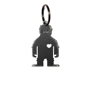 May include: A matte black metal keychain featuring a stylized creature silhouette with a textured surface. The figure has a simple face with two eyes and a mouth, a small circular cutout on the right side, and a heart-shaped cutout on the left chest.