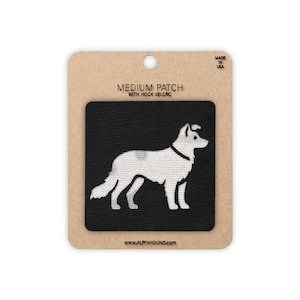 May include: A black square patch featuring a white embroidered silhouette of a dog in profile. The patch is mounted on a tan cardboard backing labelled MEDIUM PATCH WITH HOOK VELCRO, MADE IN USA, and the website address WWW.ALPINHOUND.COM.
