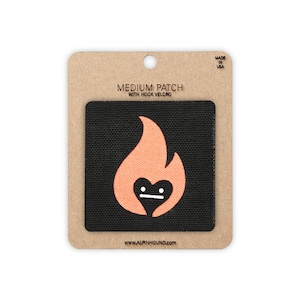 May include: A square black patch with a peach-coloured flame graphic featuring a heart-shaped centre and a flat-line face. The patch is mounted on a brown cardboard backing labelled MEDIUM PATCH WITH HOOK VELCRO and WWW.ALPINHOUND.COM, with MADE IN USA printed at the top.
