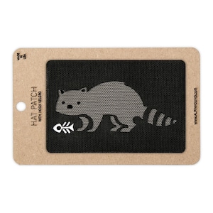 May include: A hat patch with hook and loop fastener featuring a gray raccoon holding a fish bone. The patch is on a black background and is attached to a cardboard backing. The text "HAT PATCH WITH HOOK VELCRO" is printed on the left side.