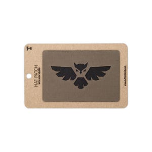 May include: A rectangular olive drab hat patch with a black embroidered owl graphic featuring spread wings. The patch is mounted on a tan cardboard display card with the text HAT PATCH WITH HOOK VELCRO, MADE IN USA, and ALPHAHOUND.COM printed.