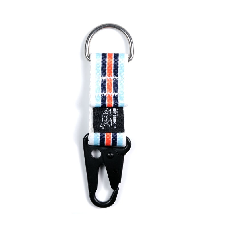 Pu&ograve; includere: Portachiavi a strisce bianche e blu con un moschettone in metallo nero e un piccolo logo nero che dice "ALPINHOUND" con un grafico di un cane che corre.