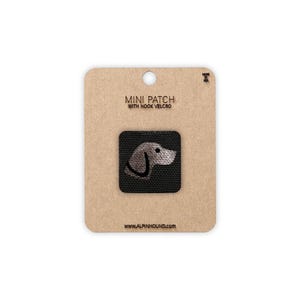 May include: A mini patch with hook velcro featuring a dog's head design. The patch is square, with a black background and a light gray dog illustration. The patch is attached to a brown cardboard backing with the text "MINI PATCH WITH HOOK VELCRO" and "WWW.ALPINHOUND.COM".