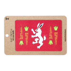 Peut inclure: Un écusson rectangulaire rouge pour chapeau avec un dos en velcro, présentant une silhouette de lapin blanc au centre, entourée de deux carottes orange avec des fanes vertes de chaque côté, fixé sur un support en carton brun.