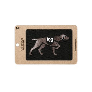 Peut inclure: Un écusson rectangulaire noir avec une silhouette de chien argentée et le texte K9 sur un support en carton beige. Le support porte le texte HAT PATCH WITH HOOK VELCRO et www.alpinhound.com. L'écusson est orienté horizontalement.