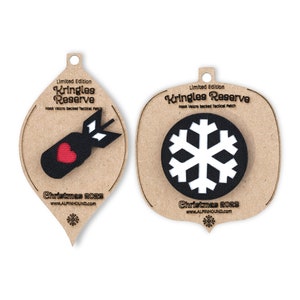 May include: Two Christmas ornaments shaped like teardrops with a brown background. The ornament on the left features a black bomb with a red heart and a white arrow. The ornament on the right features a black snowflake with a white center. Both ornaments have the text "Kringles Reserve" and "Christmas 2023" printed on them.