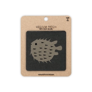 May include: A square black patch featuring a tan pufferfish graphic with small dark spikes. The patch is mounted on a rectangular tan cardboard backing labeled MEDIUM PATCH WITH HOOK VELCRO, MADE IN USA, and the website URL ALPINHOUND.COM.