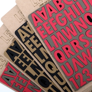 May include: Close-up of four sheets of laser cut letters. The top sheet has large, bright pink letters on a gray background. The letters are A, B, C, D, E, F, G, H, I, J, K, L, M, N, O, P, Q, R, S, T, U, V, W, X, Y, Z. The other sheets have black letters on a brown background. The letters are A, B, C, D, E, F, G, H, I, J, K, L, M, N, O, P, Q, R, S, T, U, V, W, X, Y, Z.