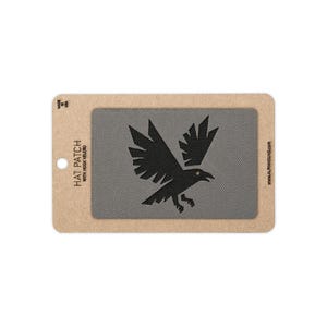May include: A rectangular gray hat patch with a black graphic of a flying raven. The patch is mounted on a tan cardboard backing card with the text HAT PATCH WITH HOOK VELCRO, MADE IN USA, and ALPHAHOUND.COM printed on the edges.