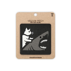 May include: A medium-sized, black patch with hook velcro. The patch features a white cat riding a gray shark. The patch is on a brown cardboard backing with the text "MEDIUM PATCH WITH HOOK VELCRO" and "MADE IN USA". The website address www.alpinhound.com is also visible.