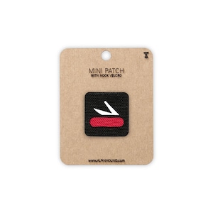 May include: A small, square black patch with a red stripe and a white graphic of a knife. The patch is attached to a cardboard backing with the text "MINI PATCH WITH HOOK VELCRO" and "WWW.ALPINHOUND.COM".