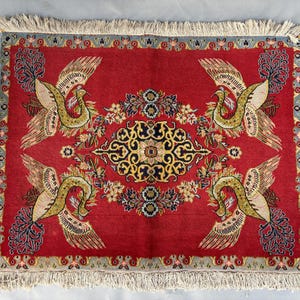Op de afbeelding: Een rechthoekig rood tapijt met een centraal bloemmotief en vier gestileerde vogelmotieven. Het tapijt heeft een crèmekleurige franje en een rand met bloemen en geometrische patronen. De vogels zijn in goud-, groen- en crèmetinten. Afmetingen ca. 100 cm x 70 cm.