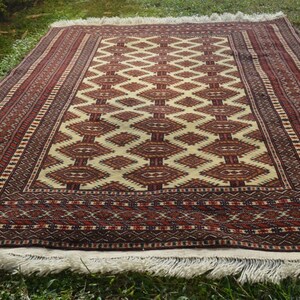 May include: A traditional rug with a cream background and a geometric pattern in shades of red, brown, and black. The rug has a fringe border.