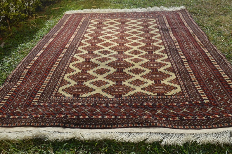May include: A traditional rug with a cream background and a geometric pattern in shades of red, brown, and black. The rug has a fringe border.