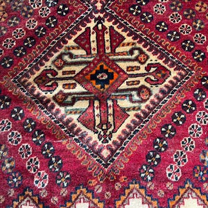 Op de afbeelding: Close-up van een vintage tapijt met een centraal ruitpatroon. Het tapijt heeft een dieprode achtergrond met ingewikkelde geometrische ontwerpen in crème, marineblauw en oranje. Bloemenmotieven en randen dragen bij aan de details.