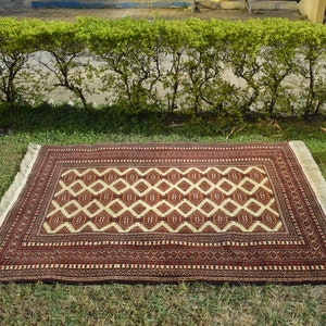 May include: A red and cream patterned rug with fringe on all sides. The rug is laid out on a green grassy lawn.