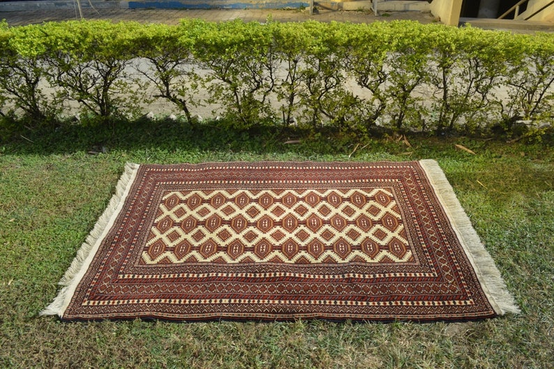 May include: A red and cream patterned rug with fringe on all sides. The rug is laid out on a green grassy lawn.
