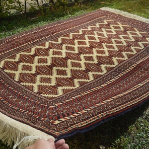 May include: A traditional hand-woven rug with a cream background and intricate red, brown, and black geometric patterns. The rug has a fringe border.