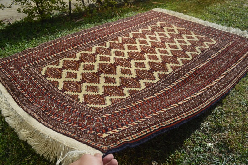 May include: A traditional hand-woven rug with a cream background and intricate red, brown, and black geometric patterns. The rug has a fringe border.