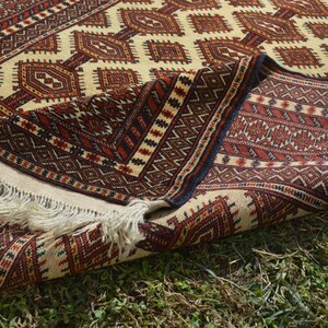 May include: A traditional rug with a cream background and intricate geometric patterns in shades of red, brown, and black. The rug is folded in half, revealing the fringe on the edge.