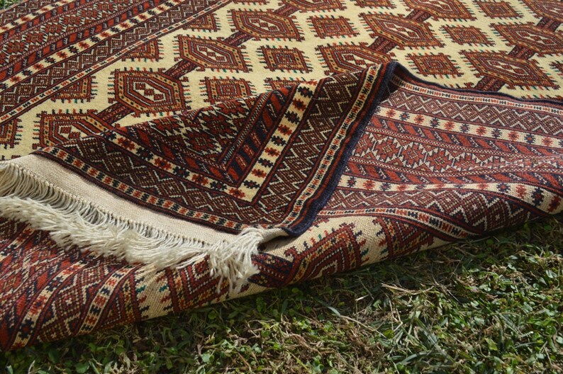 May include: A traditional rug with a cream background and intricate geometric patterns in shades of red, brown, and black. The rug is folded in half, revealing the fringe on the edge.