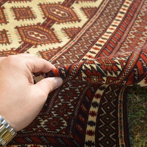 May include: A hand holding the edge of a red, white, and black patterned rug. The rug has a geometric design and is folded in half.