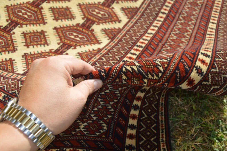 May include: A hand holding the edge of a red, white, and black patterned rug. The rug has a geometric design and is folded in half.