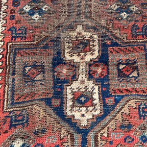 May include: Close-up of a vintage rug with a geometric pattern. The rug features a central design in navy blue, surrounded by red and beige borders. The rug's texture is visible, showcasing the woven fibres and intricate details.