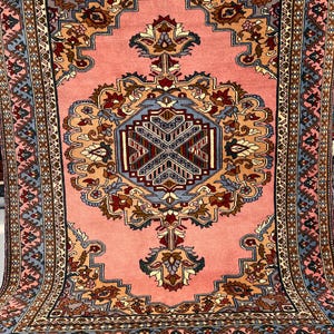 May include: A large, rectangular rug with a central medallion design in shades of peach, blue, and brown. The rug features intricate floral and geometric patterns, with a detailed border. The rug is displayed with a slight curve, showcasing its texture and design.