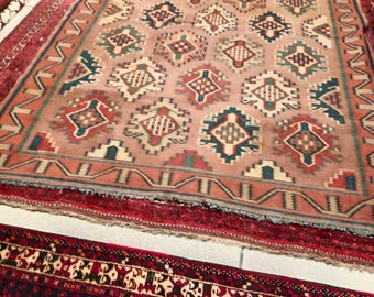 May include: A large, rectangular rug with a central pattern of repeating diamond shapes in shades of peach, teal, and red. The rug has a red border and is surrounded by other rugs in a shop setting. The rug is likely made of wool.