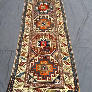 May include: A rectangular, patterned rug with a central design of geometric shapes in red, orange, blue, and cream. The rug has a cream-coloured border with repeating patterns and fringe on the ends. The rug is approximately 244 cm long.