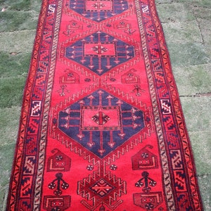 Può includere: Un tappeto rosso e nero con un design geometrico. Il tappeto ha un bordo con frange ed è fatto di lana.