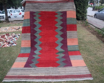 May include: A large, rectangular rug with a central red field featuring a zig-zag pattern in green and purple. The rug has a gray border and is displayed outdoors on grass. Other rugs are visible in the background.