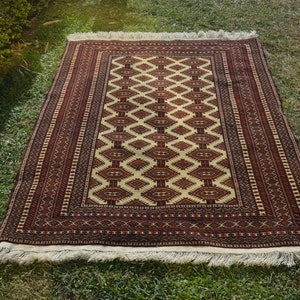 May include: A traditional rug with a cream background and a geometric pattern in shades of red and brown. The rug has a fringe border.
