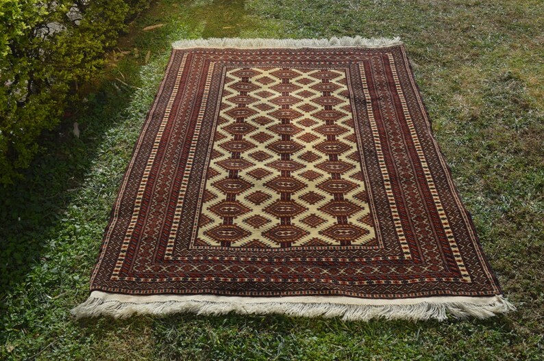 May include: A traditional rug with a cream background and a geometric pattern in shades of red and brown. The rug has a fringe border.