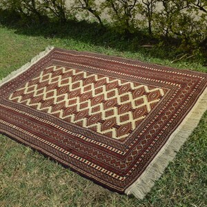 May include: A traditional rug with a geometric pattern in shades of red, white, and brown. The rug has a fringe border and is laid out on a green grassy surface.