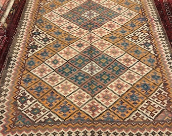 May include: A rectangular woven rug featuring a repeating diamond pattern in shades of teal, mustard yellow, and cream. The border displays intricate geometric motifs in brown and orange, finished with white fringe along the short edges.