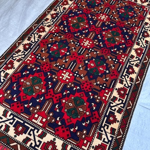 Peut inclure: Un tapis rectangulaire avec un motif géométrique détaillé. Le tapis présente un champ central rouge, bleu marine et marron, avec des accents de vert et de crème. La bordure est crème avec des formes géométriques et un bord extérieur marron.