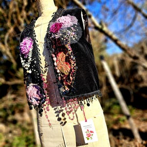 K&ouml;nnte beinhalten: Eine schwarze Samtweste mit floralen Verzierungen, Perlen und Pailletten. Die Weste hat rosa und lila Stoffblumen und baumelnde Perlenakzente. Ein Anh&auml;nger mit einer Rosenillustration h&auml;ngt an der Weste.