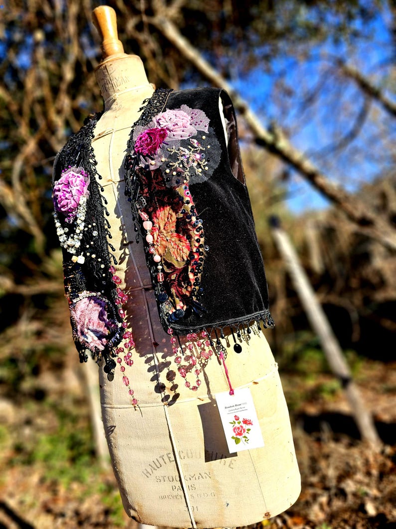 K&ouml;nnte beinhalten: Eine schwarze Samtweste mit floralen Verzierungen, Perlen und Pailletten. Die Weste hat rosa und lila Stoffblumen und baumelnde Perlenakzente. Ein Anh&auml;nger mit einer Rosenillustration h&auml;ngt an der Weste.