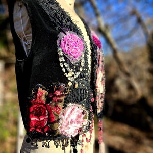 K&ouml;nnte beinhalten: Schwarze Samtweste mit floralen Aufn&auml;hern in Rot- und Rosat&ouml;nen. Die Weste hat schwarze Spitzendetails, Perlen und baumelnde Perlenfransen. Ein Kleidungsst&uuml;ck im Vintage-Stil mit einer Boh&egrave;me-&Auml;sthetik.
