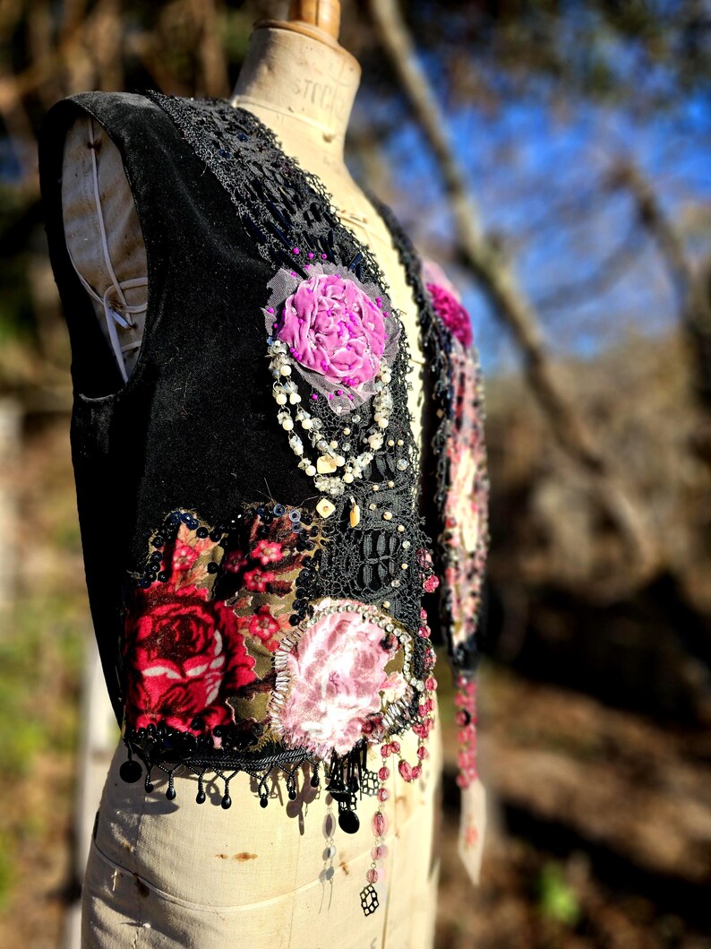 K&ouml;nnte beinhalten: Schwarze Samtweste mit floralen Aufn&auml;hern in Rot- und Rosat&ouml;nen. Die Weste hat schwarze Spitzendetails, Perlen und baumelnde Perlenfransen. Ein Kleidungsst&uuml;ck im Vintage-Stil mit einer Boh&egrave;me-&Auml;sthetik.